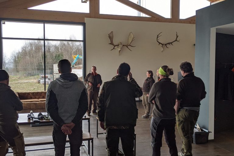 Eine Gruppe grün gekleidete Leute hören einem Jagdtrainer zu. An der Wand Hirschgeweihe.