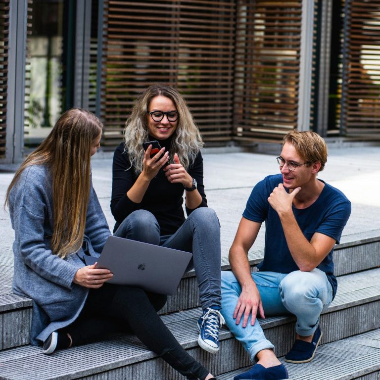 Vor einem modernen Lehrgebäude sitzen zwei junge Frauen und ein junger Mann mit Brille.